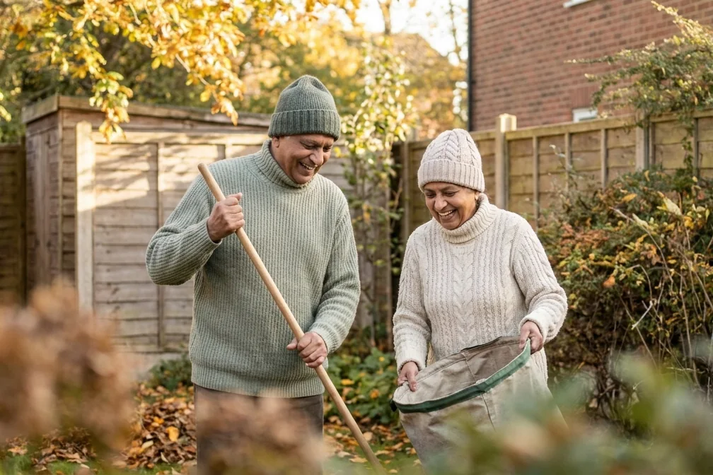 An older couple laugh together as they rake autumn leaves into a garden bag in their back garden.
