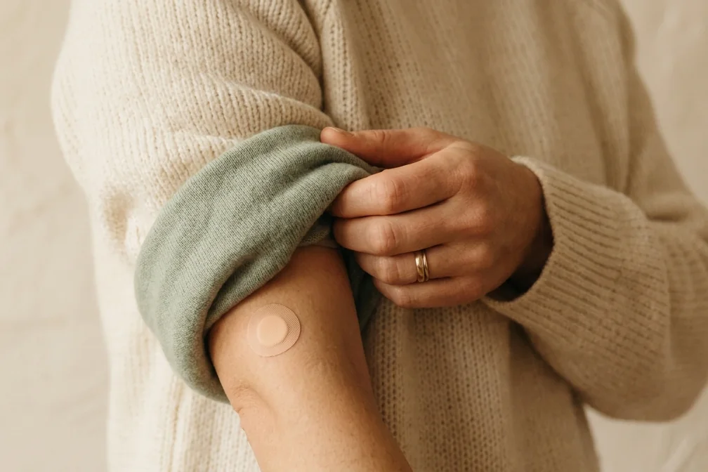 A hand rolls a knitted sleeve back down over a small round plaster on an upper arm.